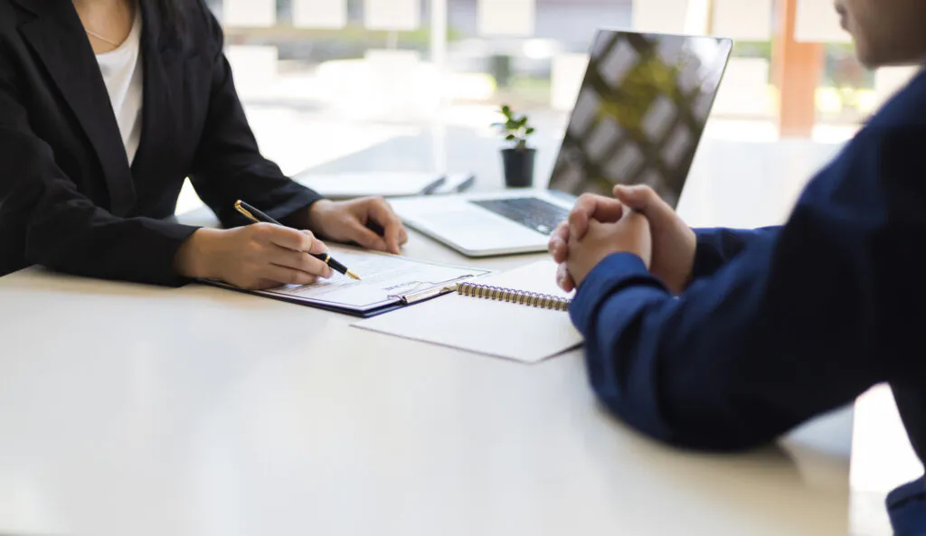 Attorney meeting with a client across a desk, reviewing and signing documents beside an open laptop.