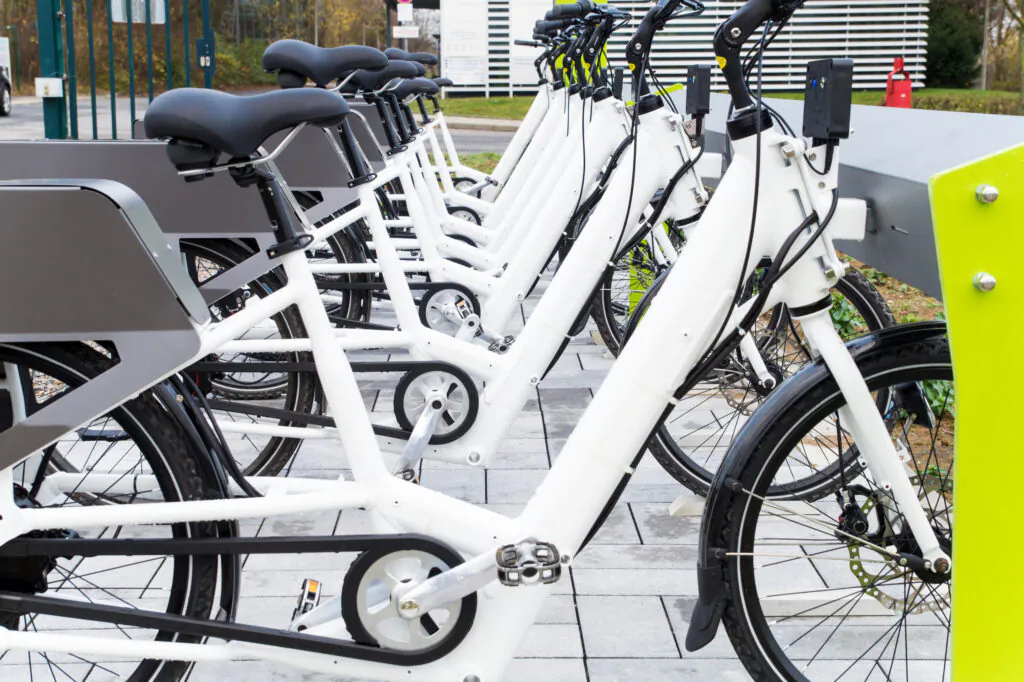 Row of white electric bicycles docked at a public bike-sharing station with charging racks outdoors.