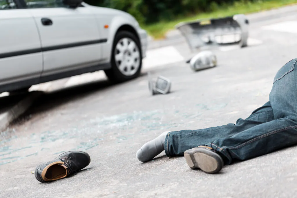 Injured pedestrian lying on the road near a car with broken vehicle parts and glass scattered nearby.