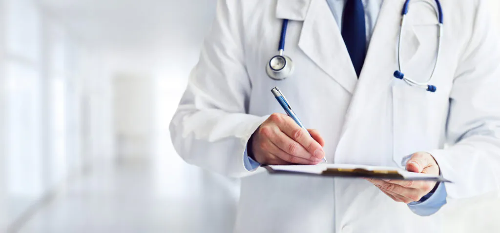 Doctor in a white coat with a stethoscope writing on a clipboard in a clinical setting.